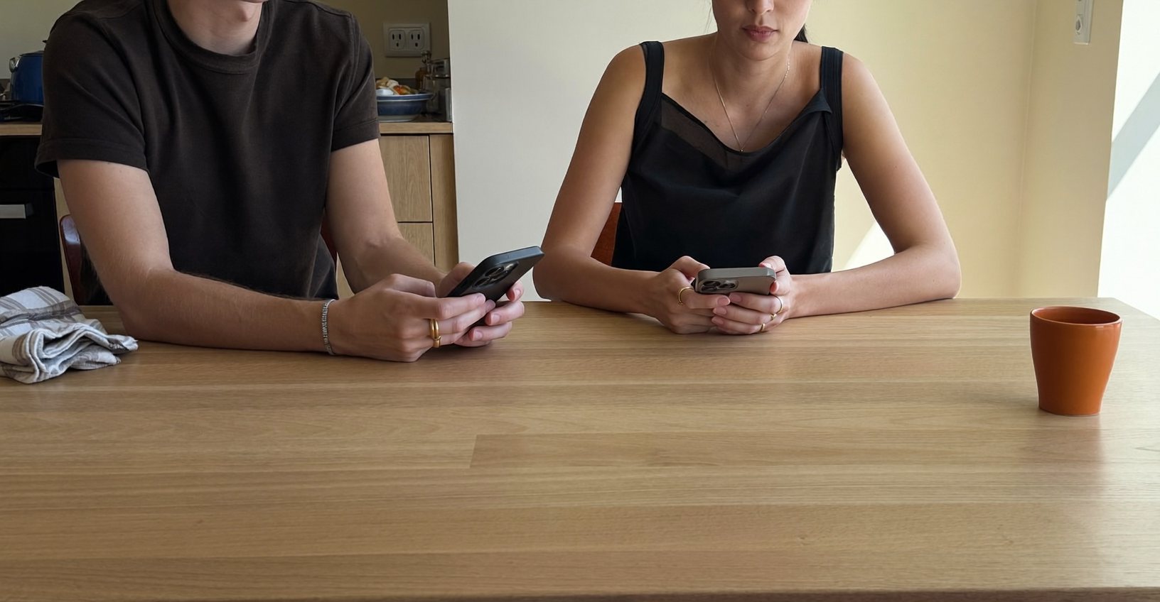 Twee mensen aan een keukentafel, elk met een telefoon, en een oranje kop